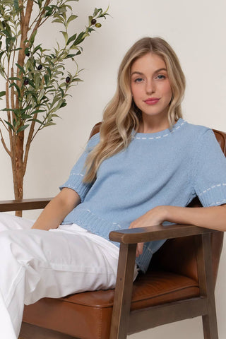 Woman sitting on a brown chair wearing a blue sweater with white text.