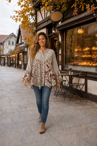 A woman walking down a cobblestone street in front of a traditional European-style building, wearing a beige and red floral print blouse with blue jeans and tan ankle boots.