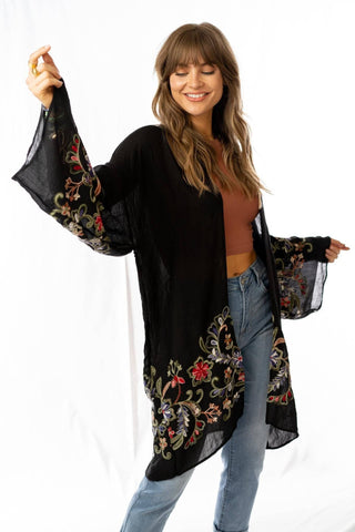 A woman modeling a black open-front cardigan with colorful floral embroidery on the sleeves and lower hem, paired with a brown crop top and blue jeans against a white studio background.