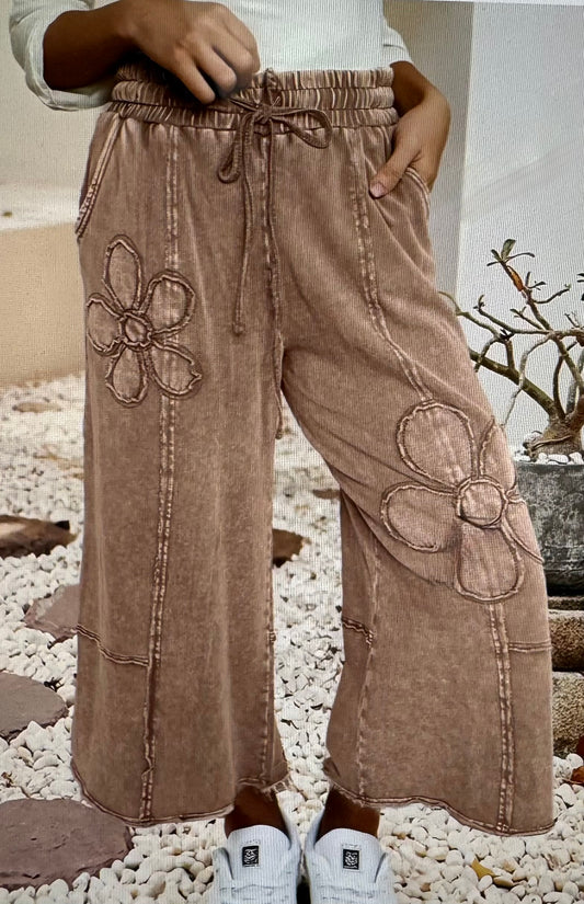 Brown wide-leg pants with floral appliqués worn by a person on a pebbled surface.