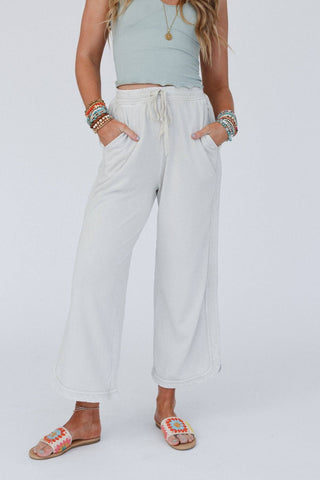A model wearing light gray wide-leg sweatpants with a drawstring waist, styled with a light blue ribbed tank top and colorful woven slide sandals against a plain white studio background.
