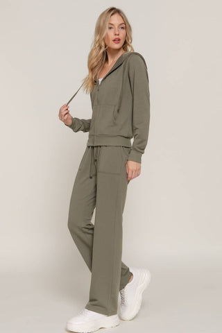 A woman modeling a matching olive green lounge set consisting of a drawstring hoodie and wide-leg sweatpants, paired with white sneakers, against a plain light gray studio background.